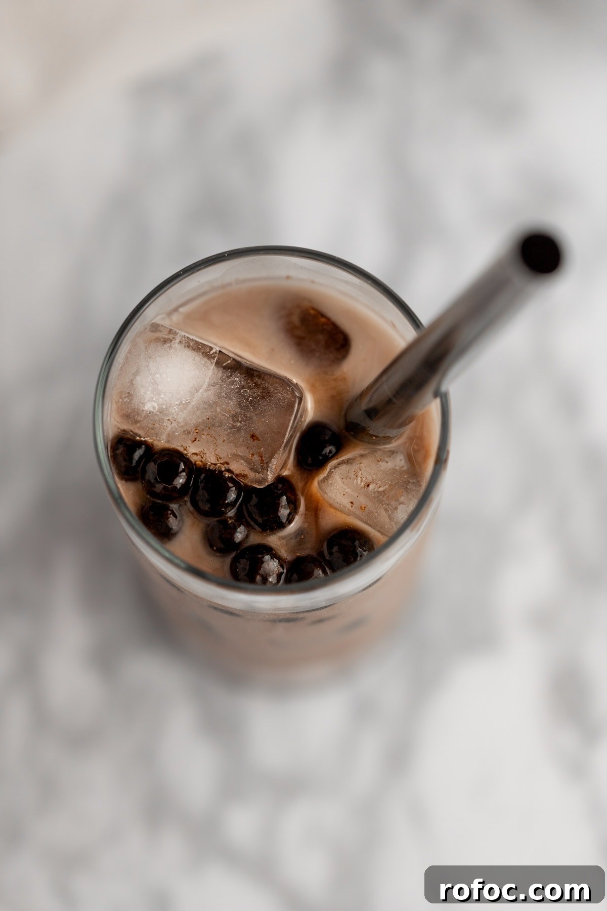 Chocolate Milk Tea Recipe: How to Make Creamy Cocoa Bubble Tea 5 Overhead photo of a glass of bubble tea, with a metal straw, ice and boba, on a grey marbled background.