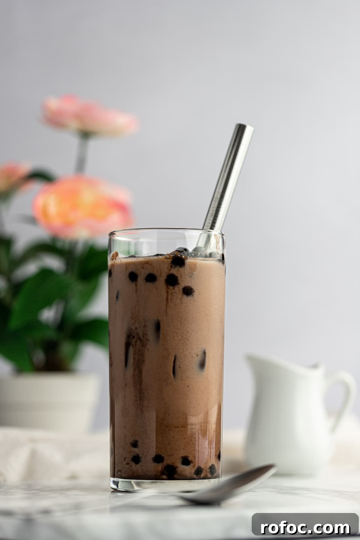 Chocolate Milk Tea Recipe: How to Make Creamy Cocoa Bubble Tea 6 Up close photo of a glass of chocolate milk tea, filled with ice and boba, with a spoon in front of the glass, and a small creamer behind, on a white background.