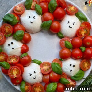 Festive Christmas Wreath Salad Recipe for Holiday Parties 3 A festive plate arranged like a holiday wreath with mozzarella balls decorated as smiling faces, surrounded by cherry tomatoes and fresh basil leaves.