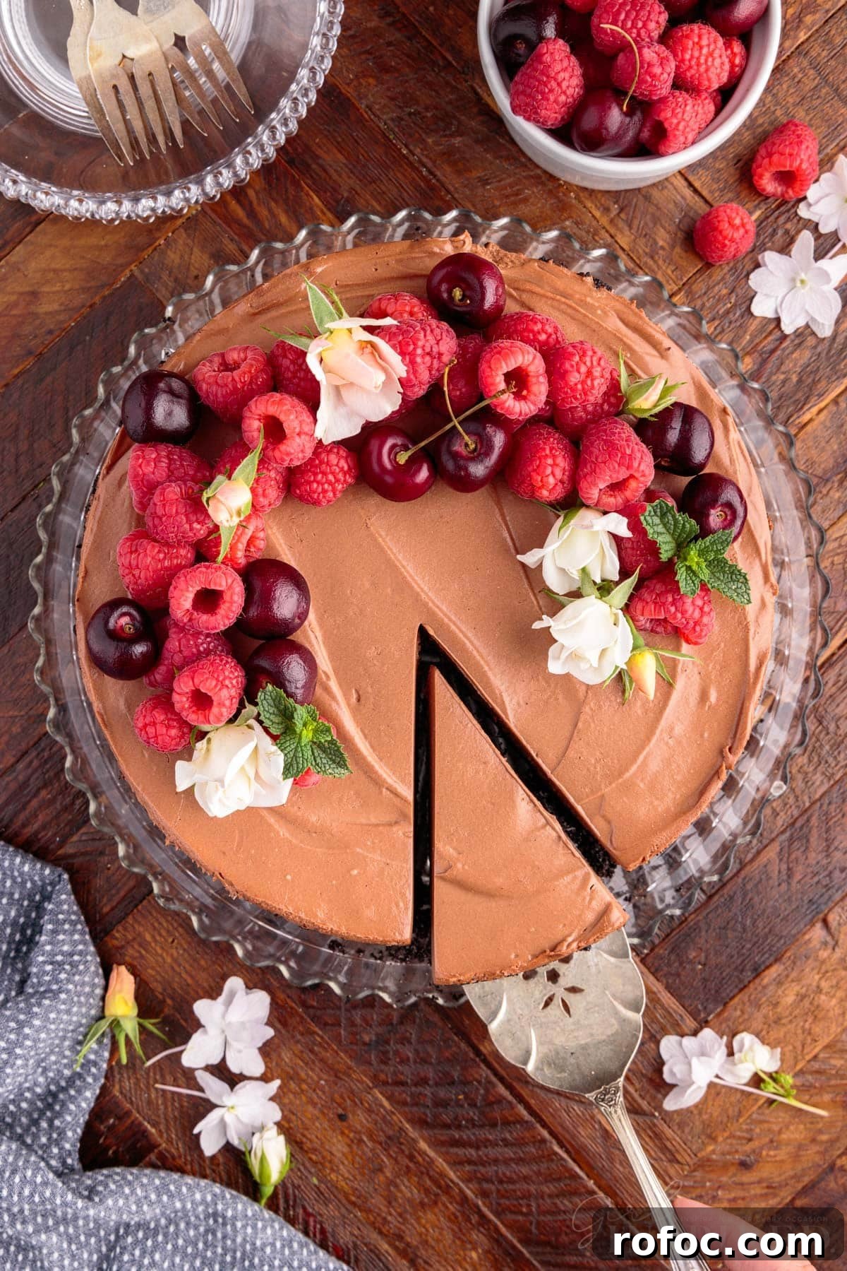 No Bake Chocolate Cheesecake garnished with flowers, cherries, raspberries, and mint leaves, with pie server lifting the first slice.