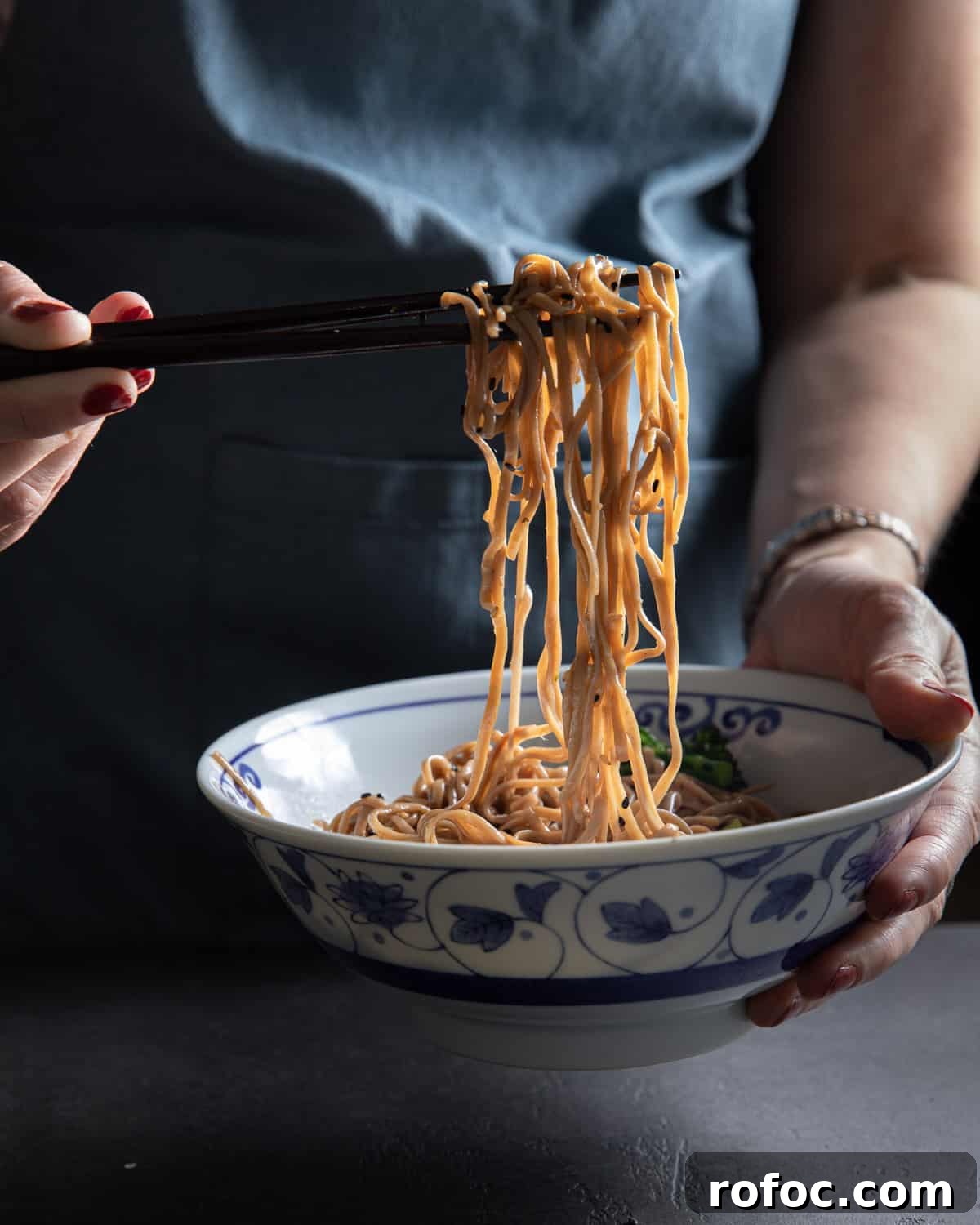 Photo of a person holding up noodles from a bowl with chopsticks.