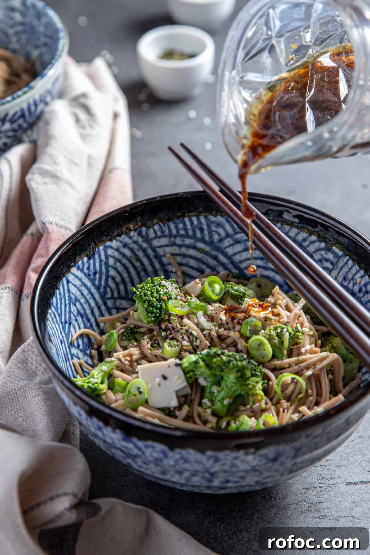 Photo of a patterned bowl of soba noodles with broccoli and the dressing being poured on top from a glass jug.