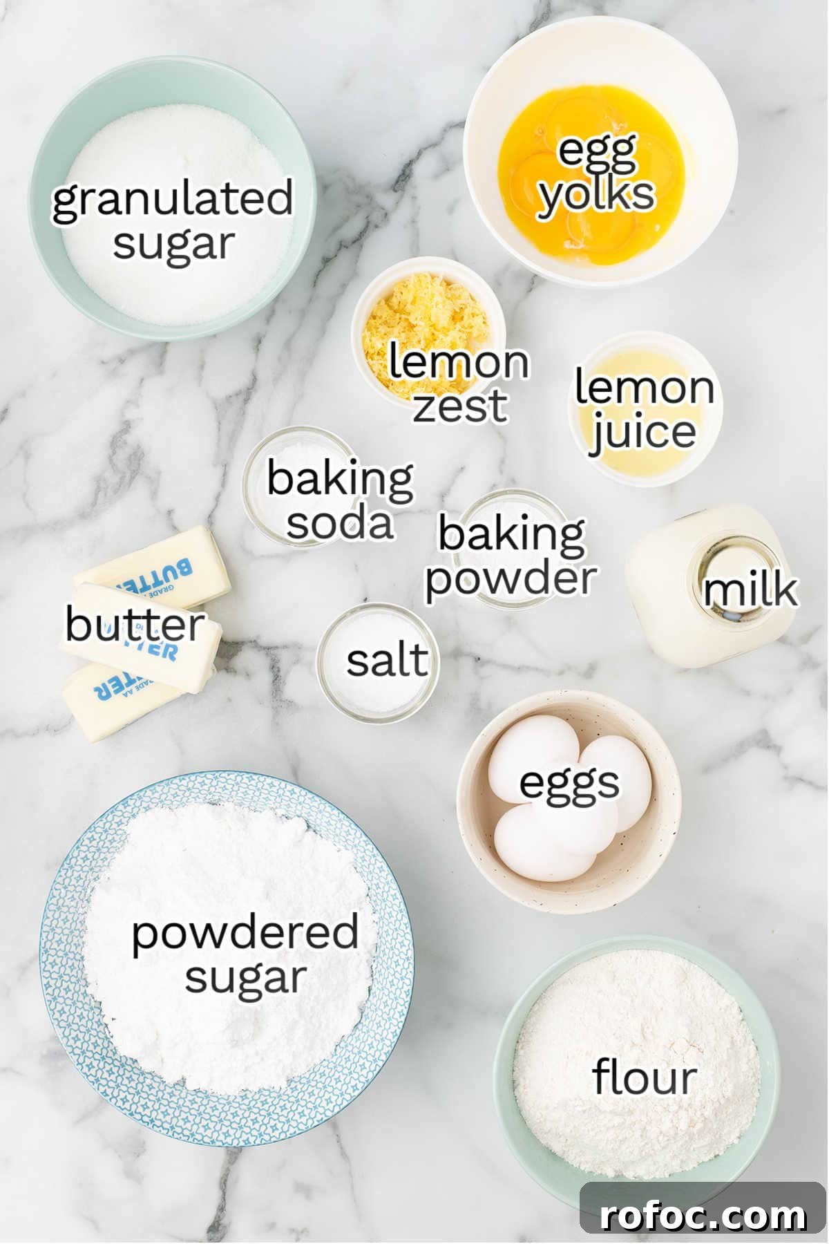 Baking ingredients for Lemon Cake on a marble countertop, including granulated sugar, powdered sugar, flour, butter, eggs, egg yolks, lemon zest, lemon juice, baking soda, baking powder, salt, and milk.