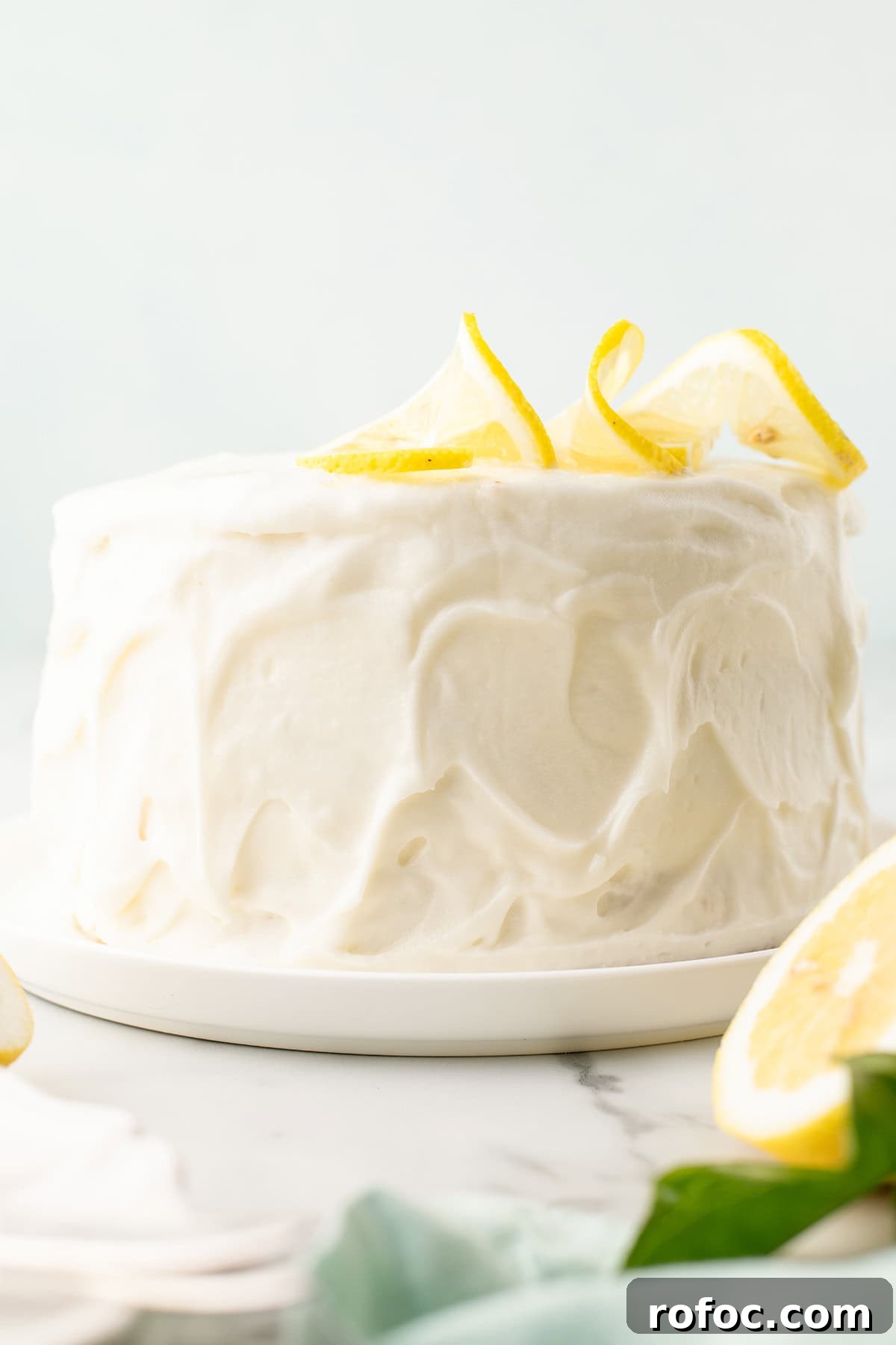 A round frosted cake sits on a white plate, decorated with lemon slices and lemon zest curls on top.