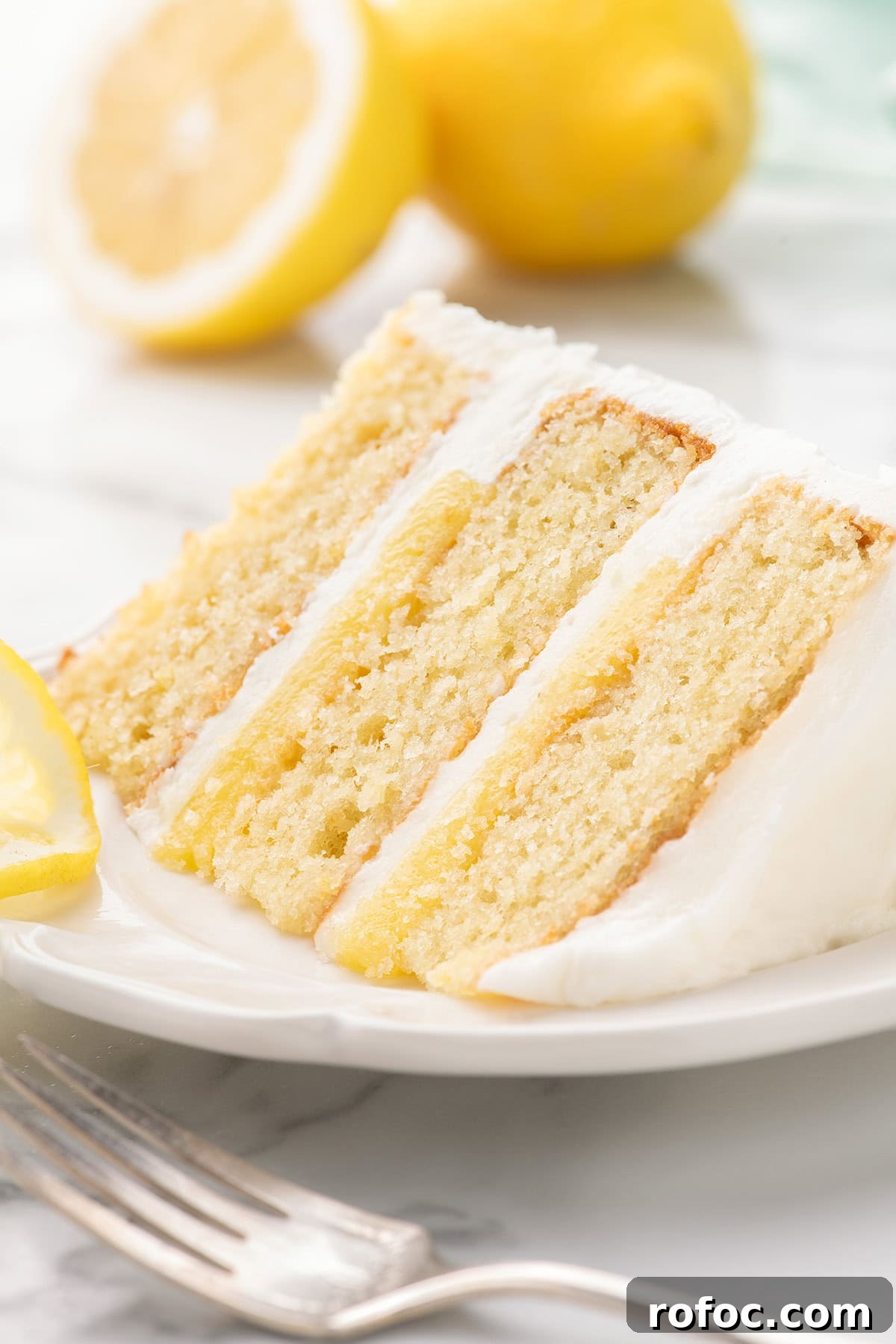A slice of three-layer lemon cake with white frosting and lemon filling on a white plate, with a fork and lemon halves in the background.