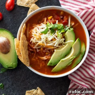 White bowl containing tortilla soup topped with shredded cheese, avocado and cilantro.