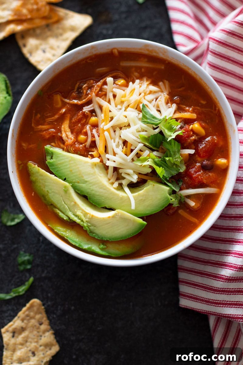 Bowl containing tortilla soup containing topped with garnishes, tortilla chips on table.