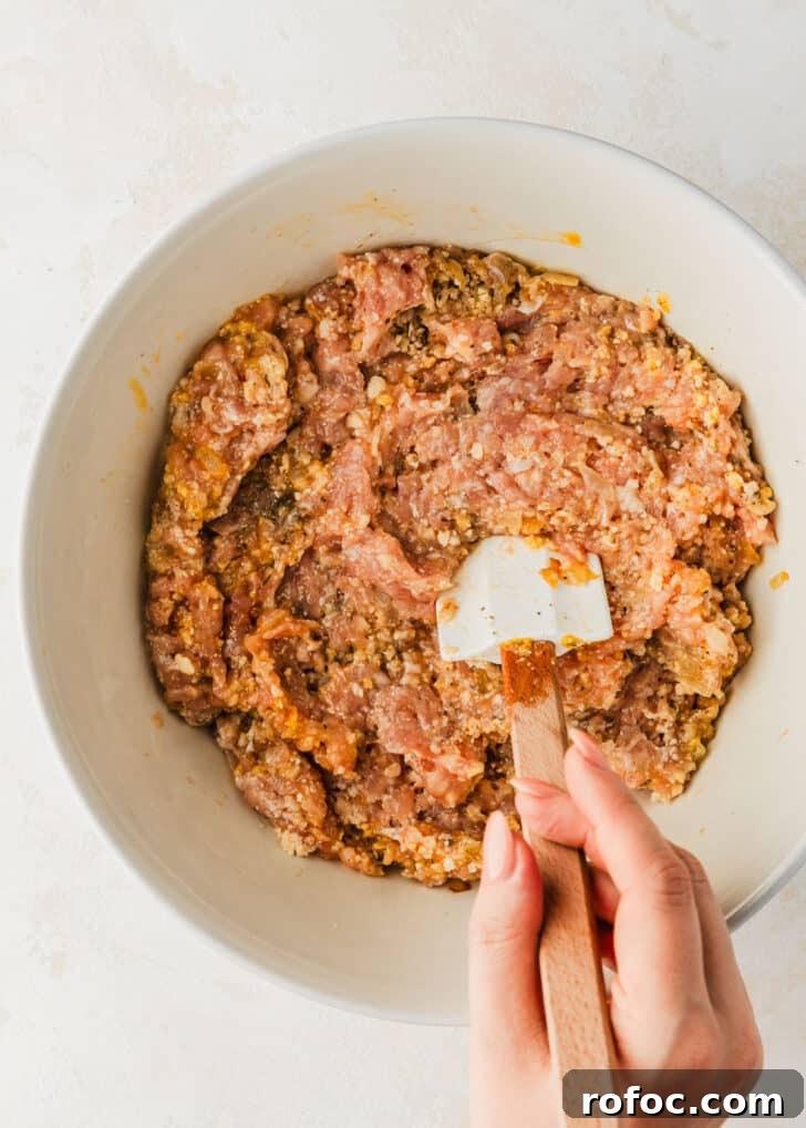 Crispy Air Fryer Chicken Meatballs Recipe 6 A hand using a rubber spatula to mix meatball mixture in a white bowl.