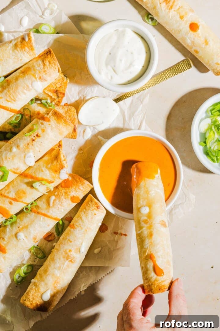 Spicy Buffalo Chicken Taquitos Recipe for Game Day Snacks 2 Baked buffalo chicken taquitos lined up on parchment paper, with a woman's hand dipping into buffalo sauce