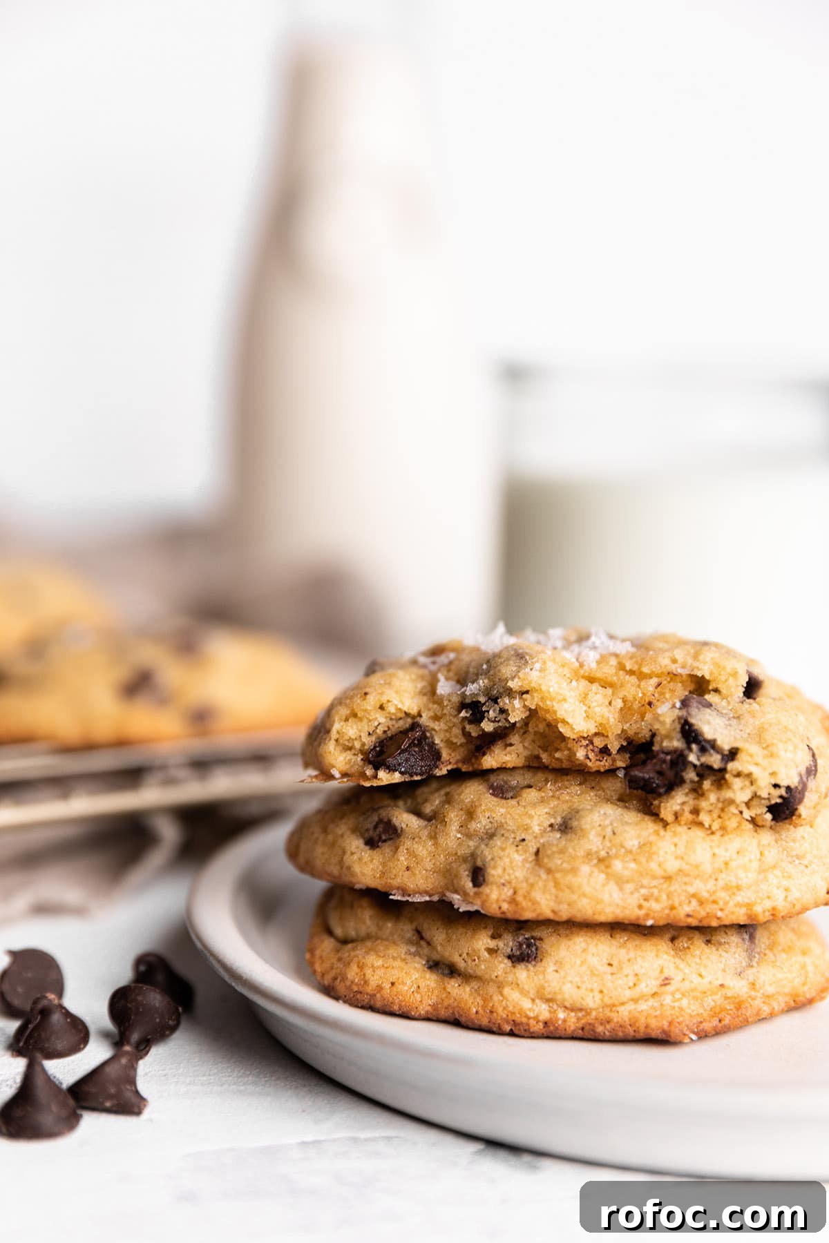 A stack of chocolate chip cookies on a plate with a glass and container of milk in the back. 