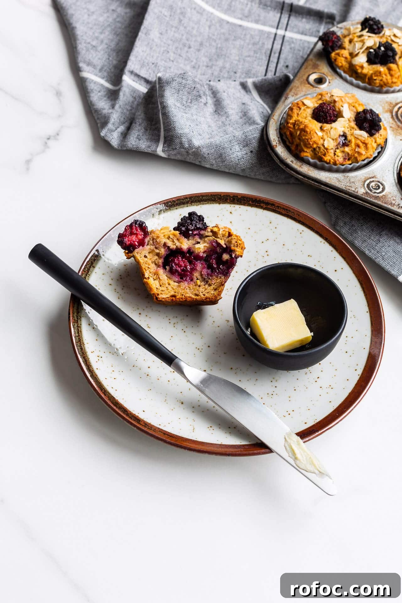 Hearty Apple and Blackberry Muffins Recipe for Fall Baking 6 Hearty oat apple muffins with blackberries on a ceramic plate with butter and a knife, vintage muffin pan in background