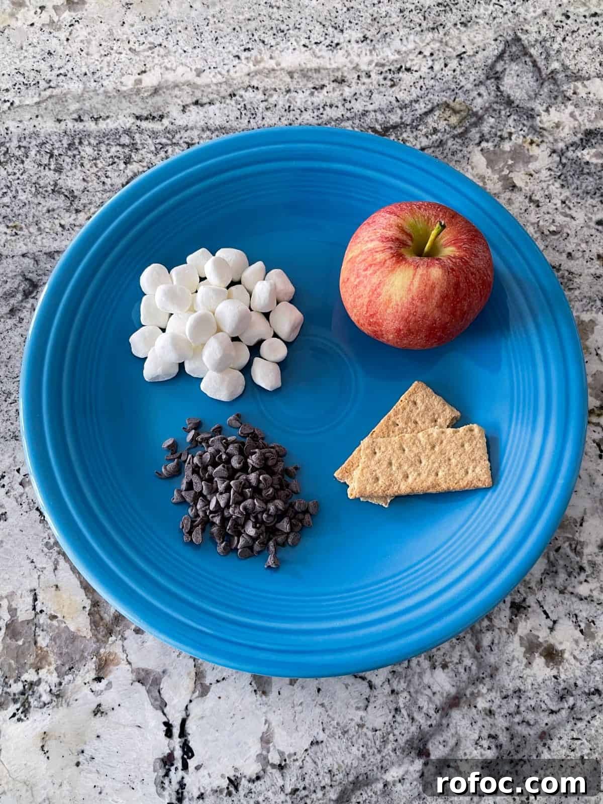Apple S'mores Nachos: Crispy Cinnamon Apple Slices with Chocolate & Marshmallow Dip 3 Mini marshmallows, chocolate chips, two graham crackers and whole apple on blue plate on granite.