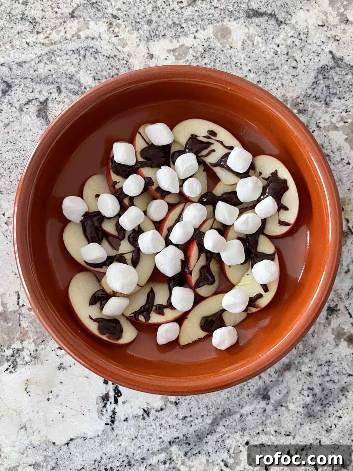 Apple S'mores Nachos: Crispy Cinnamon Apple Slices with Chocolate & Marshmallow Dip 5 Sliced apple, chocolate drizzle and mini marshmallows in orange dish on granite.