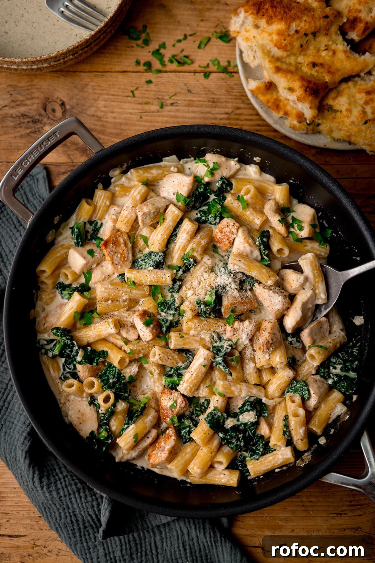 One-Pot Chicken Rigatoni Alfredo Recipe for Creamy Weeknight Meals 5 A frying pan filled with creamy rigatoni alfredo pasta, chicken pieces, and kale, topped with grated cheese and parsley. Garlic bread is on a plate nearby.