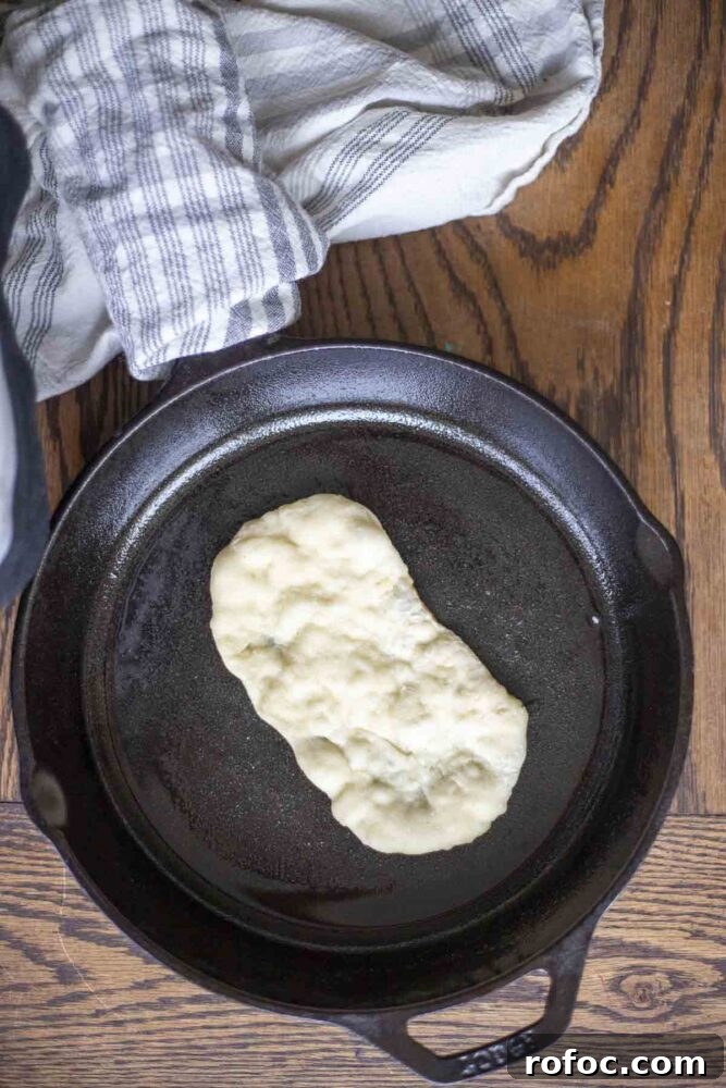 Sourdough Flatbread Recipe Using Active Starter or Discard (No Yeast) 12 sourdough flatbread cooking on a cast iron skillet on a wood countertop top with a blue stripped towel wrapped around the handle.