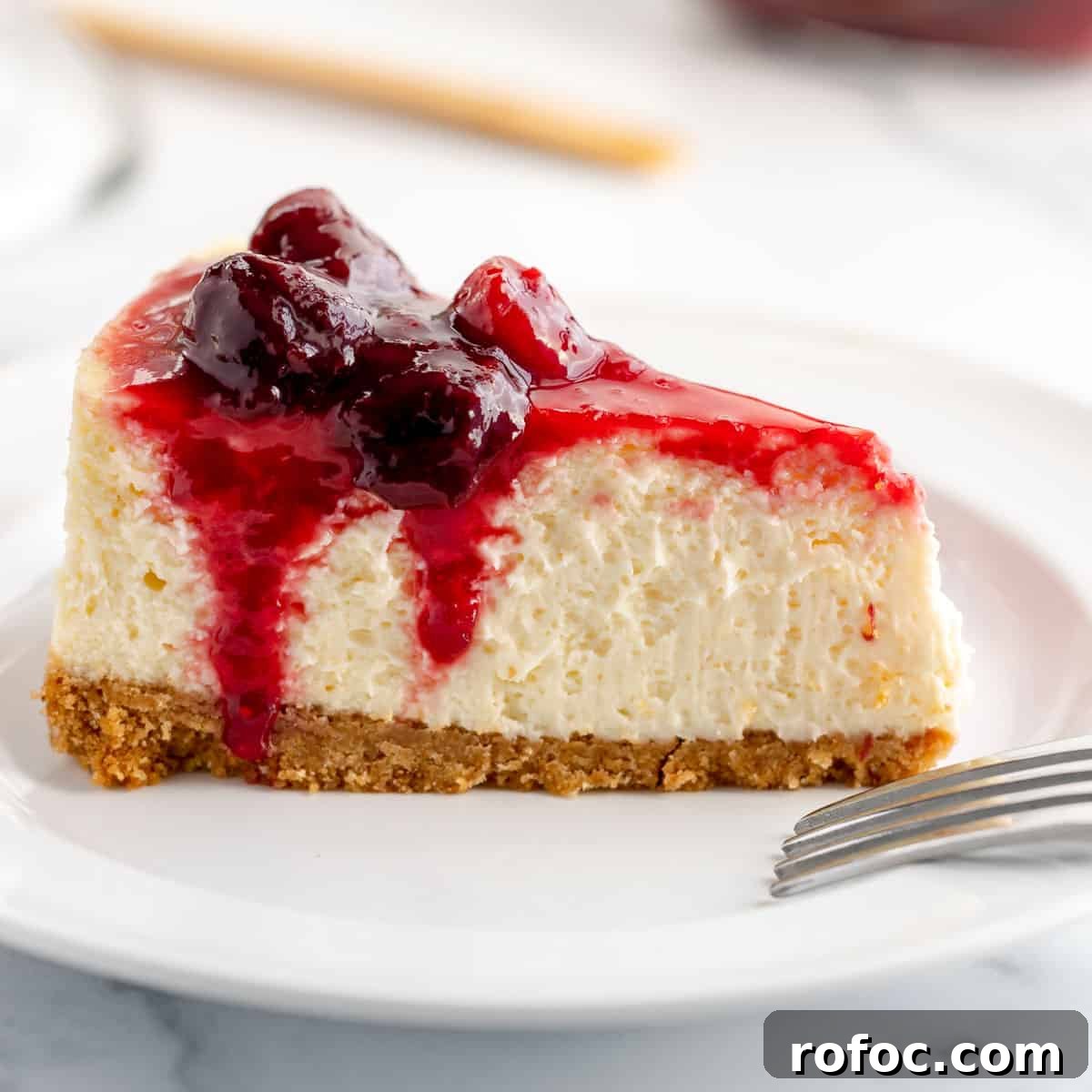 Decadent Cherry Cheesecake Recipe with a Buttery Crust 17 A slice of cheesecake with cherry sauce on top, sitting on a white plate. A fork is off to the side.