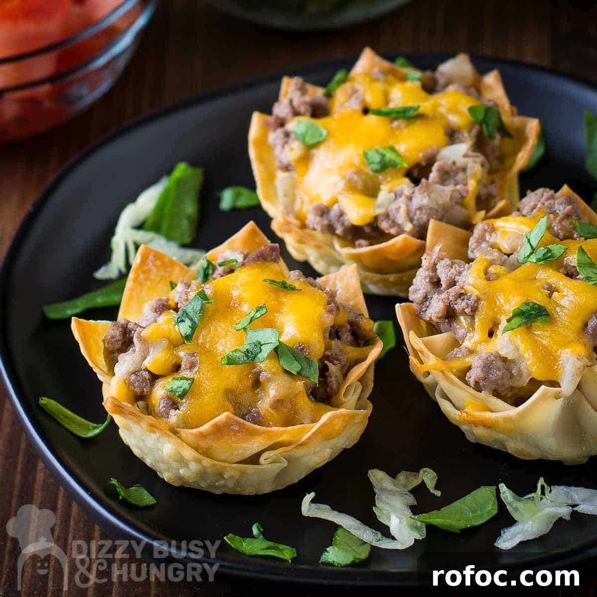 Wonton Cheeseburger Cups: Mini Cheeseburger Bites in Crispy Wrappers 6 Side shot of three wonton cheeseburger cups on a black plate garnished with herbs on a wooden surface.