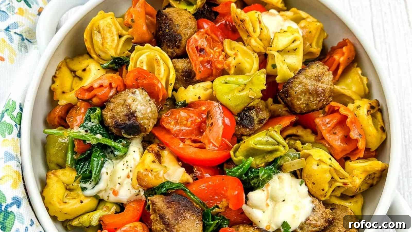 19 Fall Cookout Recipes for Sunday Dinner and Game Day 12 A close-up of a dish featuring colorful tortellini mixed with meatballs, spinach, and chopped red bell peppers, topped with melted cheese, served in a white bowl.