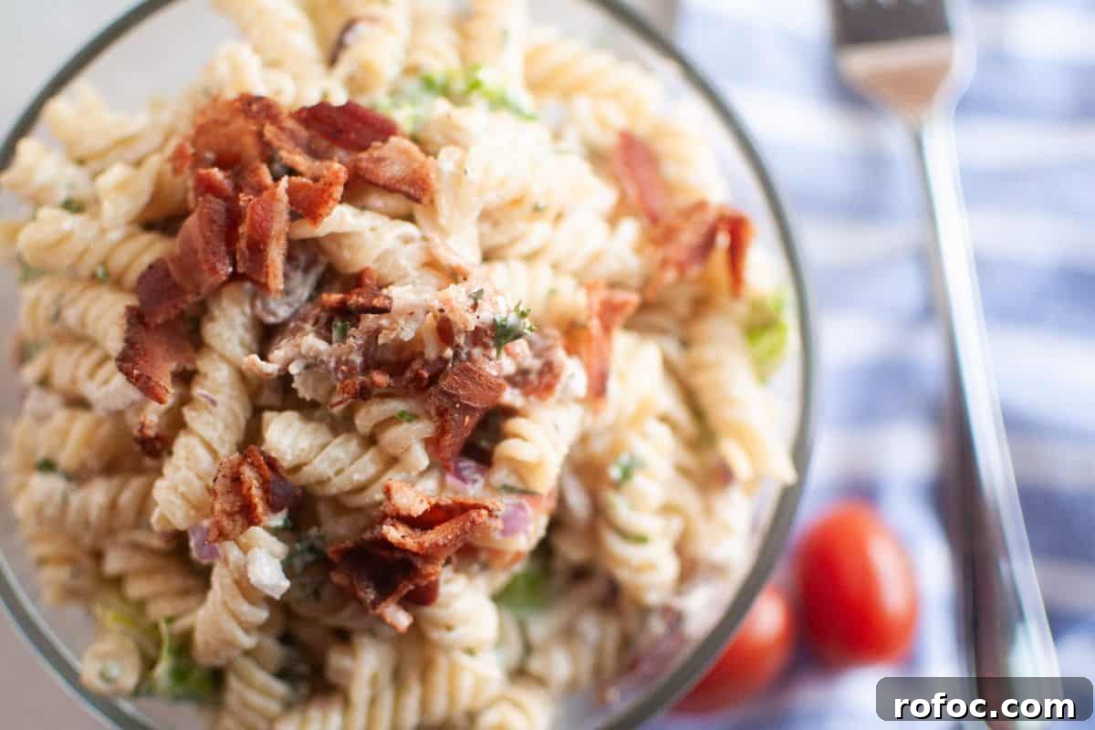19 Fall Cookout Recipes for Sunday Dinner and Game Day 19 Overhead shot of a glass bowl filled with pasta salad, featuring spiral pasta, creamy dressing, pieces of bacon, and other visible ingredients. A blue striped cloth and cherry tomatoes are visible in the background.