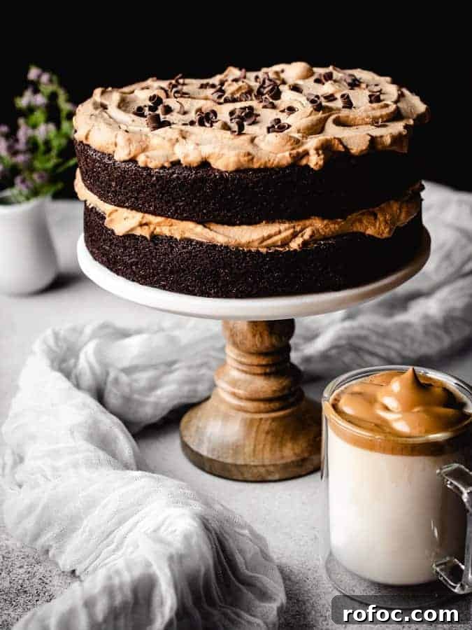 Coffee chocolate cake on a cake stand with dalgona coffee in front and flowers behind