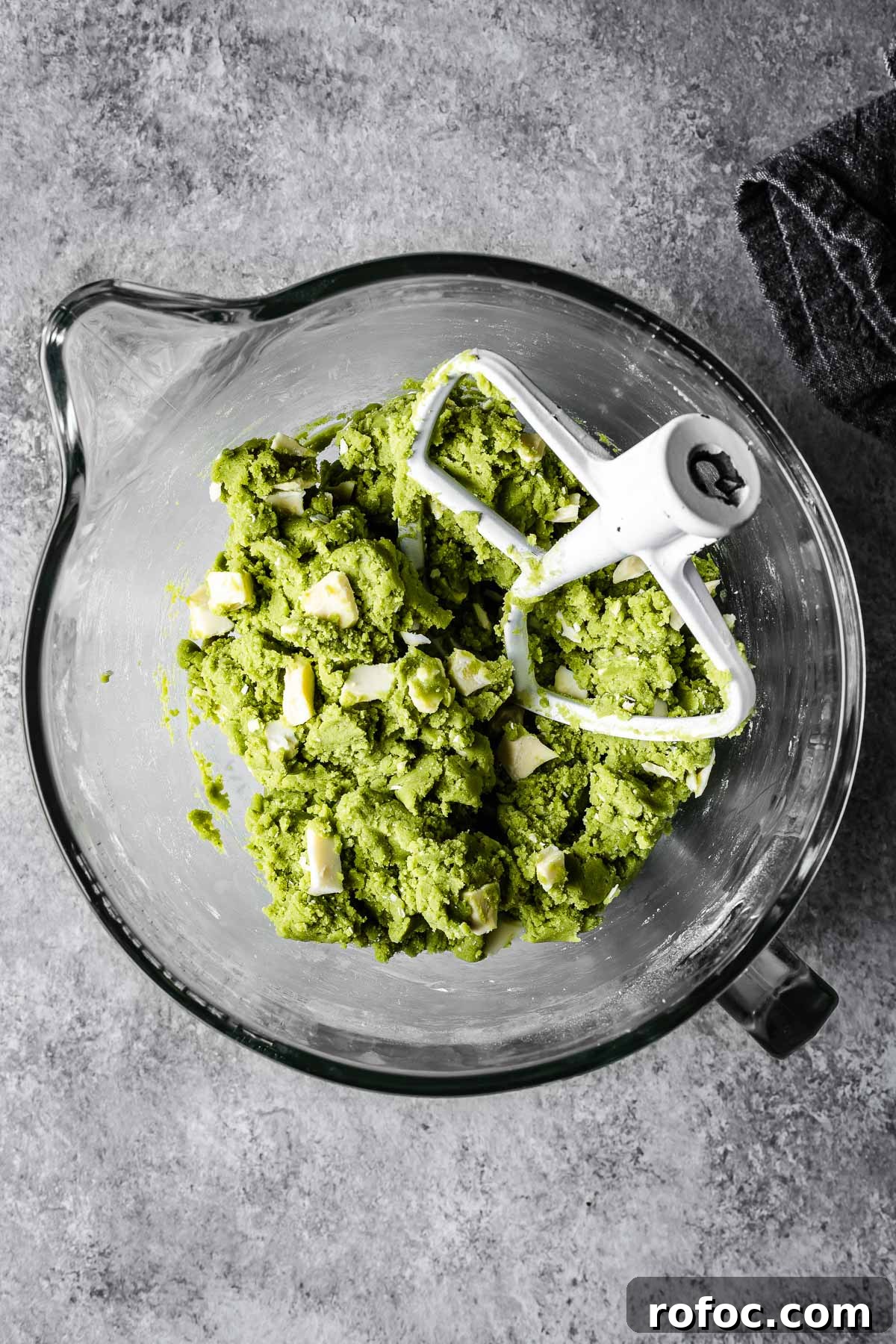 Green cookie dough in a glass mixer with white chocolate chunks added in.