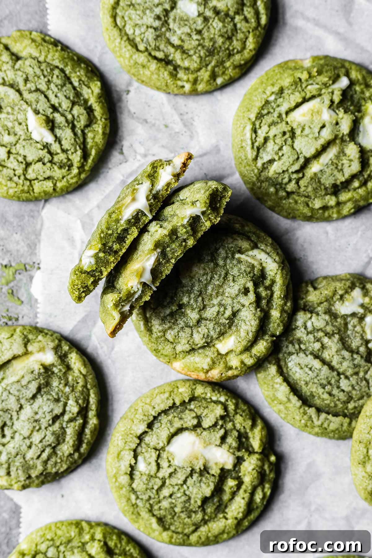 A matcha white chocolate cookie is broken in half, revealing the gooey melted chocolate inside. There are many other matcha cookies surrounding it.