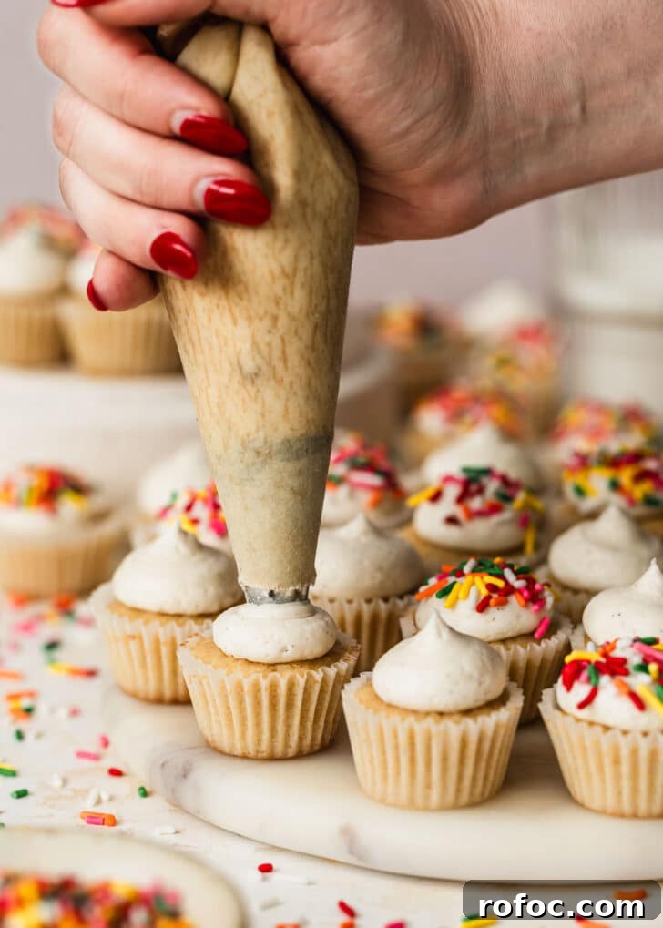 Mini Vanilla Cupcakes: Tender Bite-Sized Recipe 12 A hand decorating mini vanilla cupcakes with vanilla buttercream on a white platter next to sprinkles.