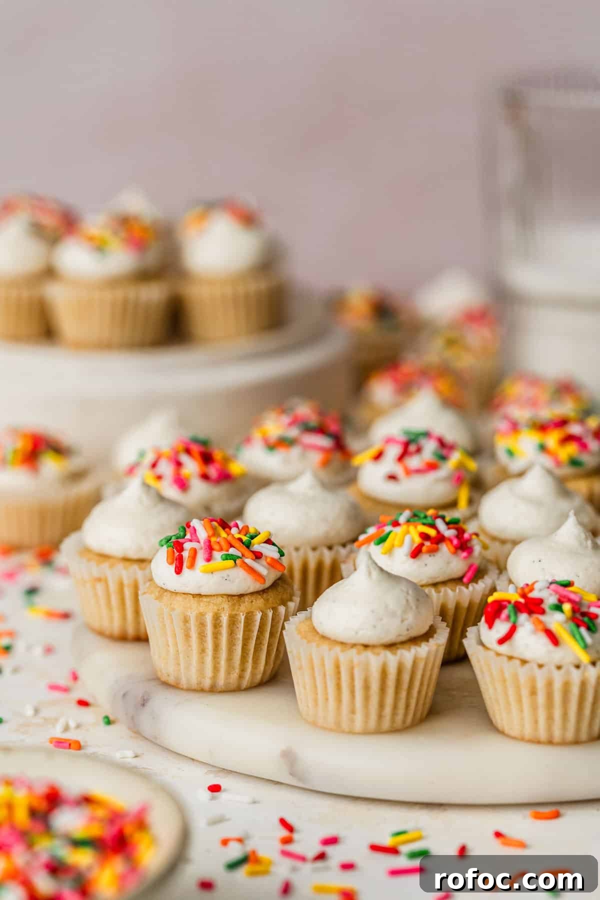 Mini Vanilla Cupcakes: Tender Bite-Sized Recipe 13 A white platter of mini vanilla cupcakes with rainbow sprinkles on a pink background next to a white bowl of sprinkles and a glass of milk.