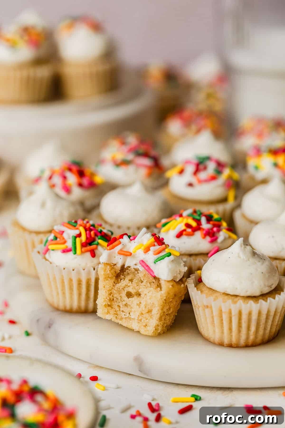 Mini Vanilla Cupcakes: Tender Bite-Sized Recipe 14 A cupcake with a bite taken out of it on a white platter next to rows of mini vanilla cupcakes, a glass of milk, and a white bowl of sprinkles with a pink backdrop.
