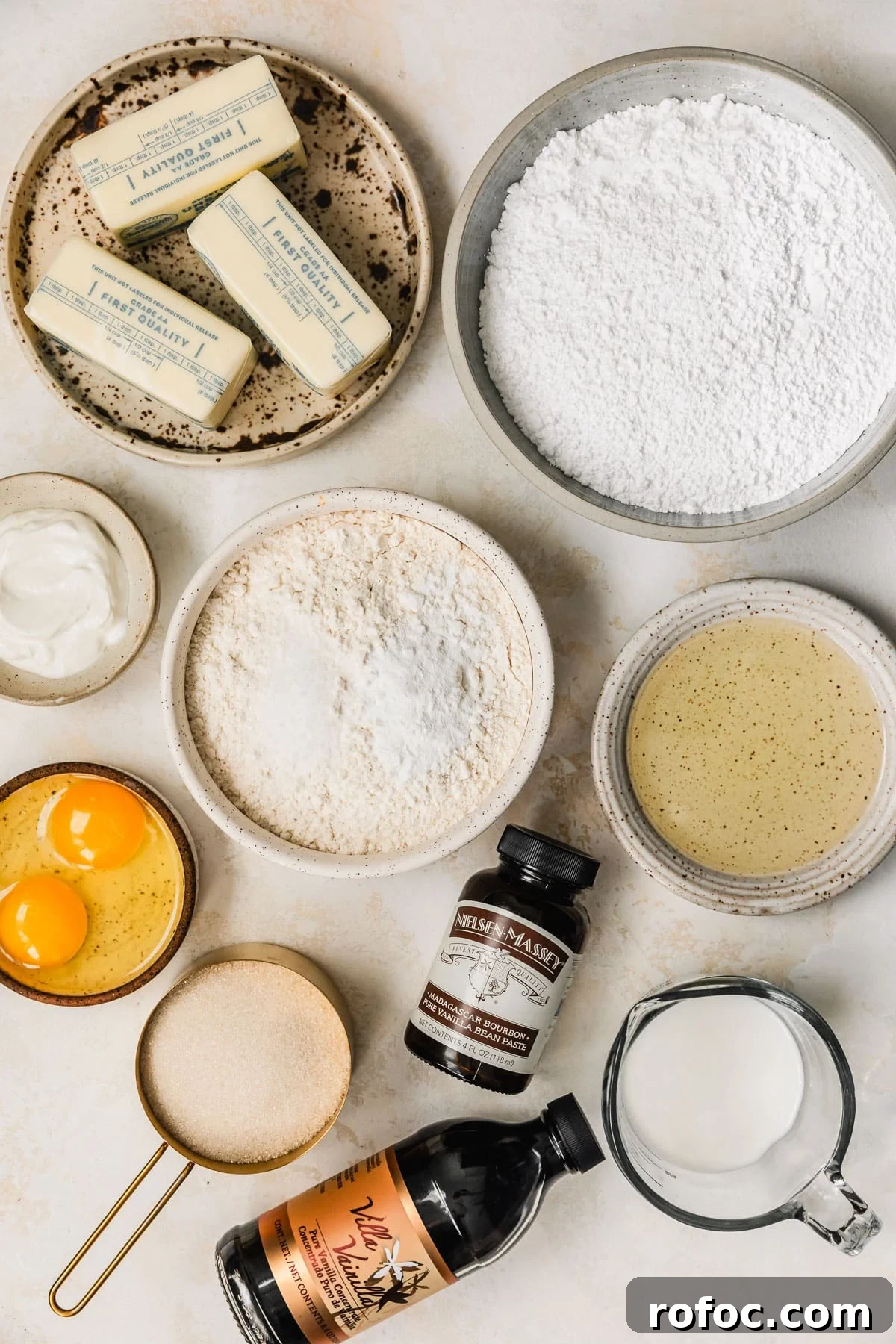 Mini Vanilla Cupcakes: Tender Bite-Sized Recipe 3 White and brown bowls of butter, flour, eggs, sour cream, powdered sugar, oil, sugar, and milk on a tan counter next to bottles of vanilla.