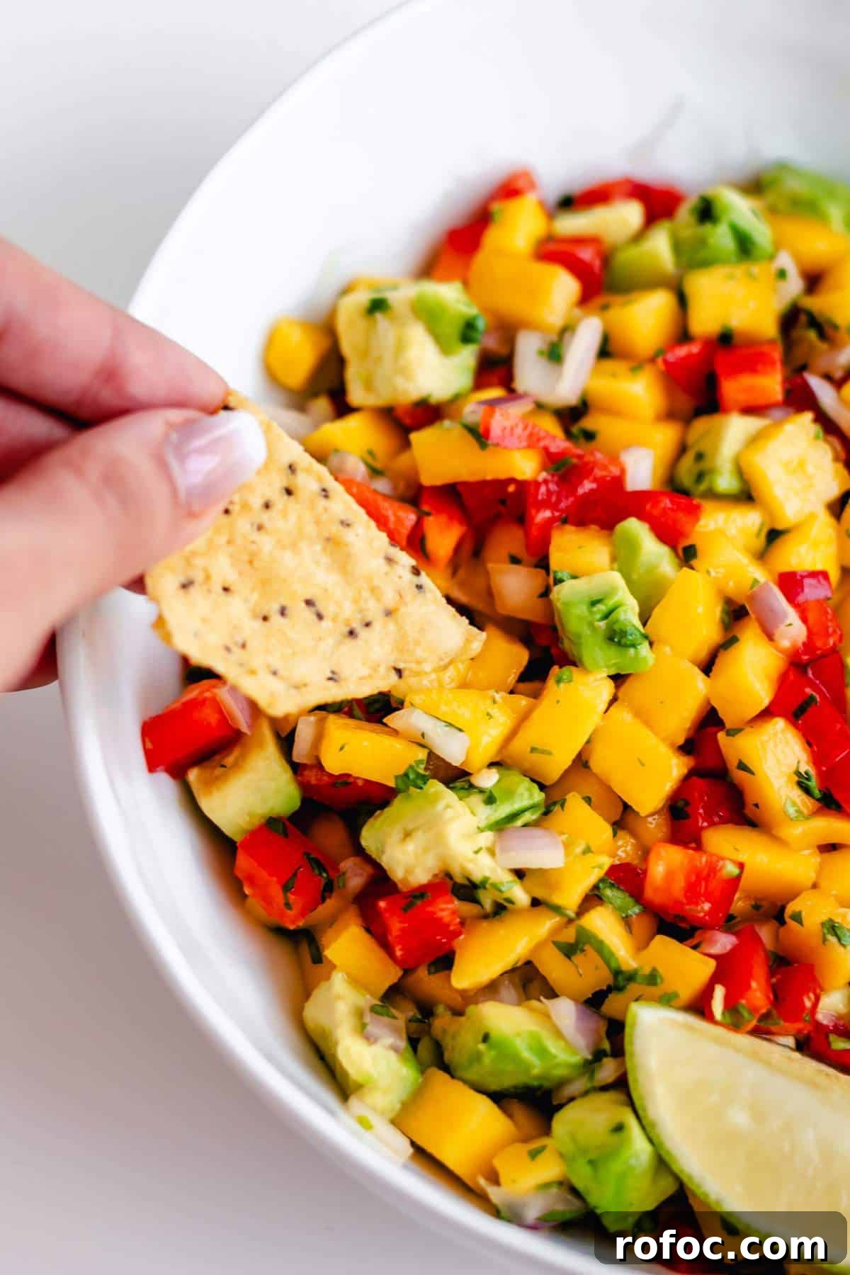 A tortilla chip scooping mango avocado salsa out of a white bowl